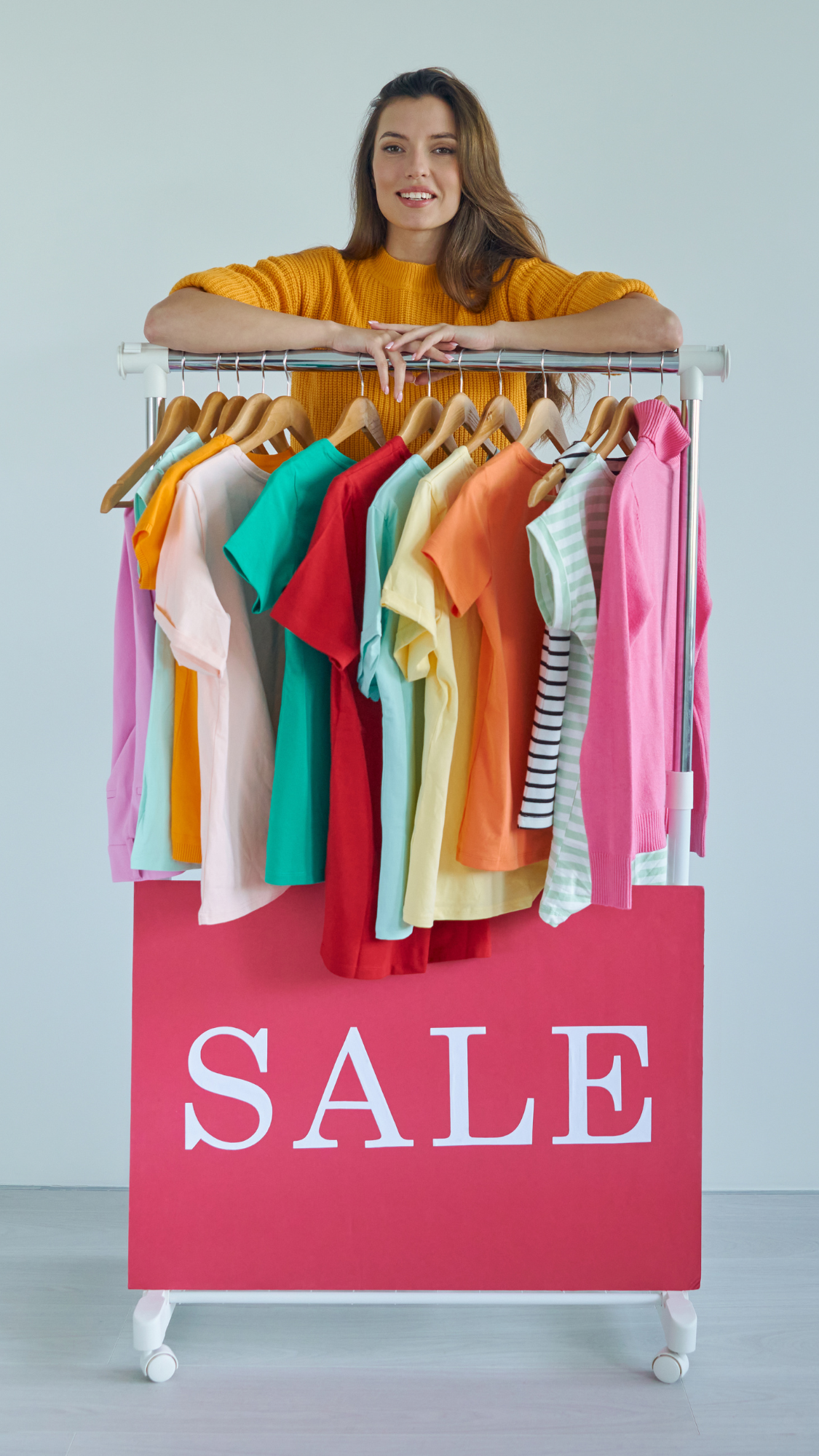 Woman standing behind a clothing rack with colorful shirts and a 'SALE' sign.